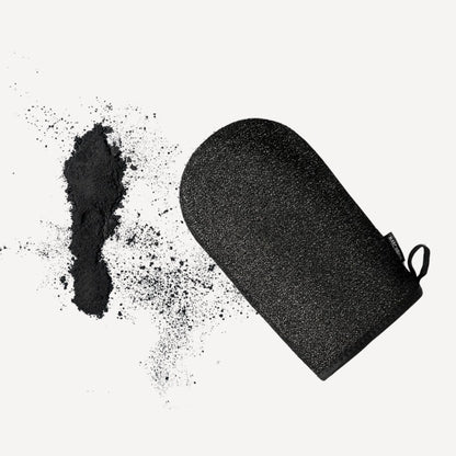 Marc Inbane Exfoliating Glove placerad på vit bakgrund med exfolierande kolpulver, visar handske för djupgående hudrengöring