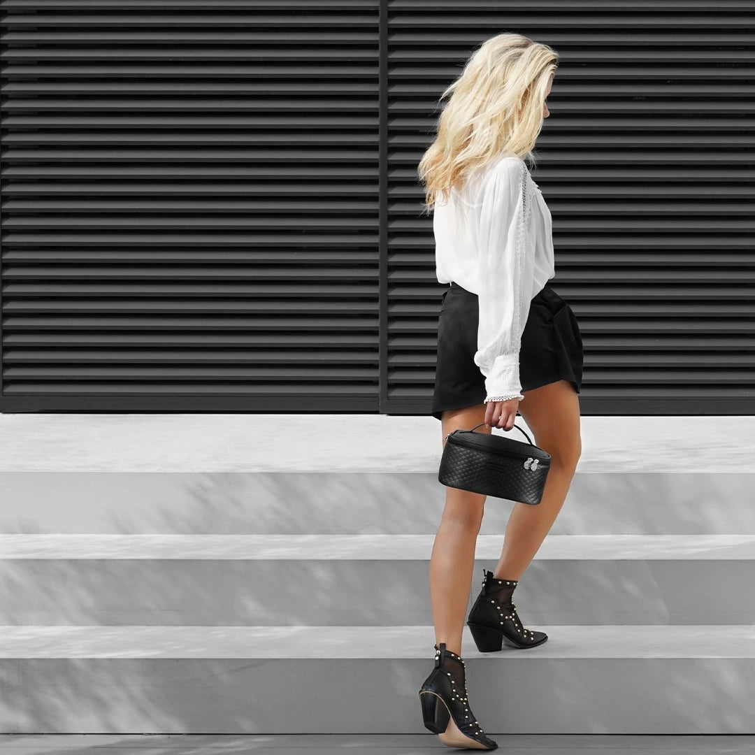 Kvinna i vit blus och svarta shorts går uppför trappa med en svart quiltad Marc Inbane kosmetikväska i handen, matchad med svarta boots med nitar.