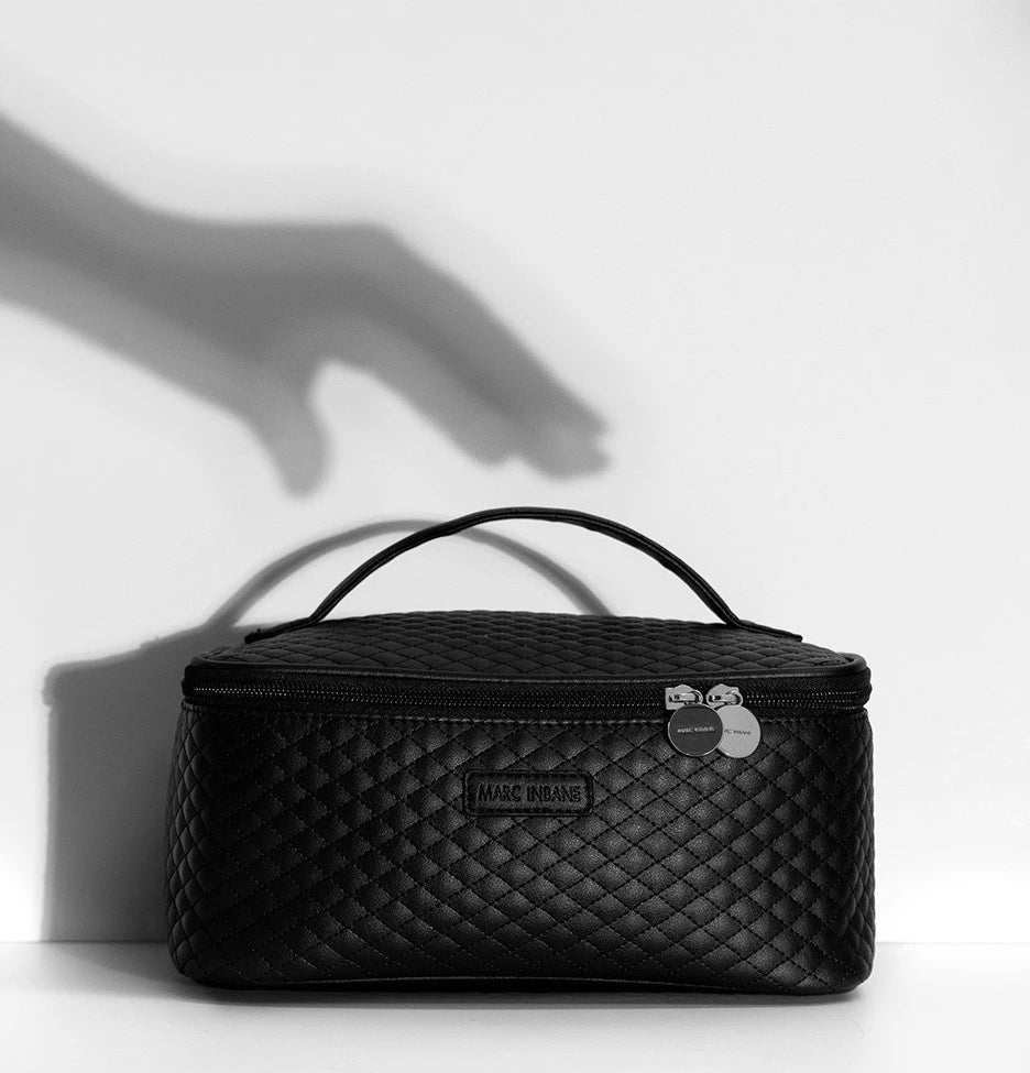 Elegant svart kosmetikväska från Marc Inbane med quiltad design och silverdetaljer, fotograferad mot vit bakgrund med skuggspel av en hand.